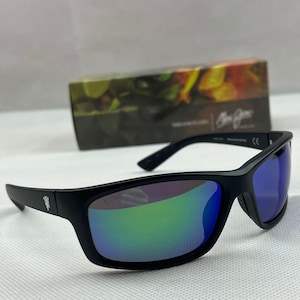 May include: Black sunglasses featuring a matte frame and reflective lenses that shift between green and blue hues. The temples display a small silver emblem, and the glasses are positioned in front of a rectangular box with a blurred floral print.