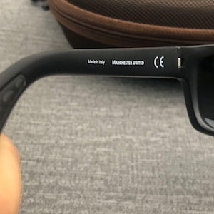 Puede incluir: Gafas de sol negras con el texto "Made in Italy" y "MANCHESTER UNITED" impreso en blanco en la patilla. Las gafas de sol tienen una marca CE y lentes oscuras.