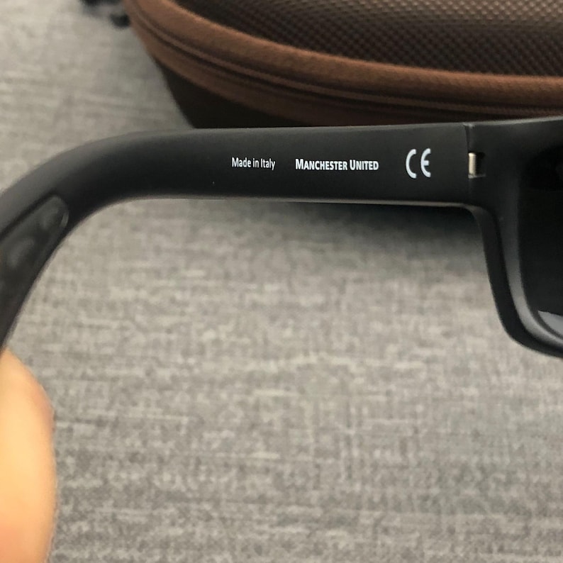Puede incluir: Gafas de sol negras con el texto "Made in Italy" y "MANCHESTER UNITED" impreso en blanco en la patilla. Las gafas de sol tienen una marca CE y lentes oscuras.
