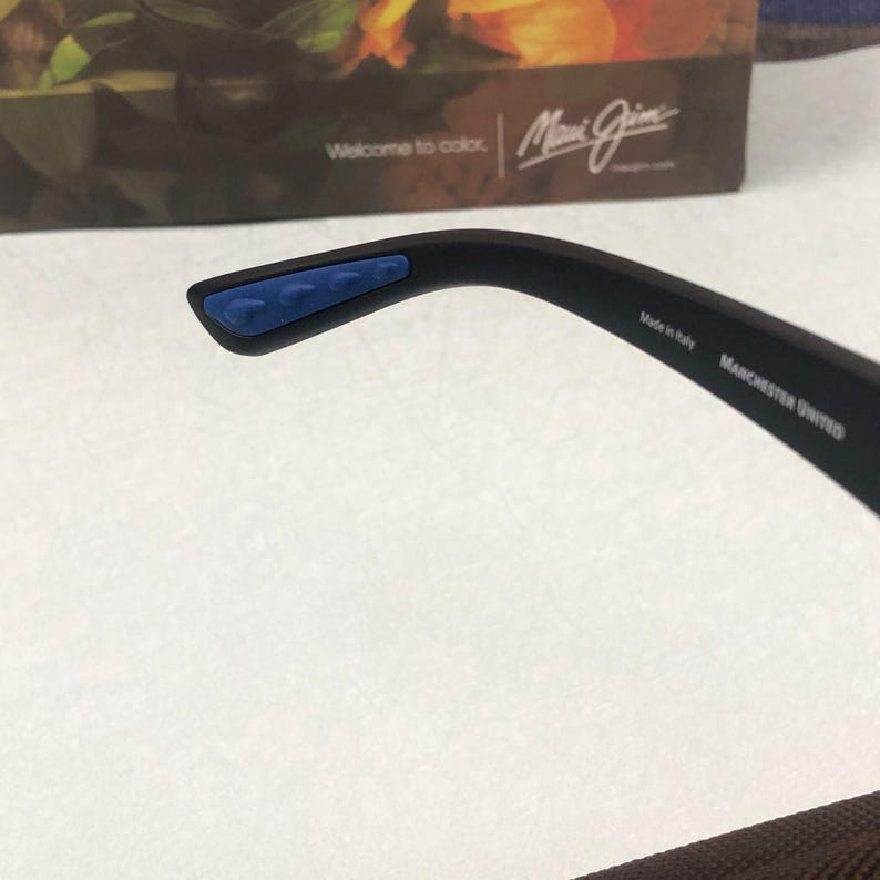 May include: Black sunglasses with blue accents on the temple tips. The text "MANCHESTER UNITED" is printed on the arm. The sunglasses are made in Italy.