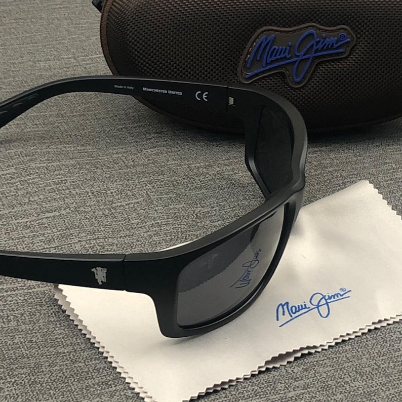 Puede incluir: Gafas de sol negras con lentes oscuras, interior blanco y el texto "MANCHESTER UNITED" en la patilla. Las gafas descansan sobre un pa&ntilde;o de limpieza blanco con el logotipo "Maui Jim". Un estuche marr&oacute;n est&aacute; en el fondo.