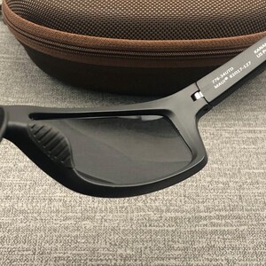 Puede incluir: Gafas de sol negras con lentes oscuras y acabado mate. Las gafas tienen forma rectangular y un agarre texturizado en el lateral. El estuche es marr&oacute;n con cremallera.