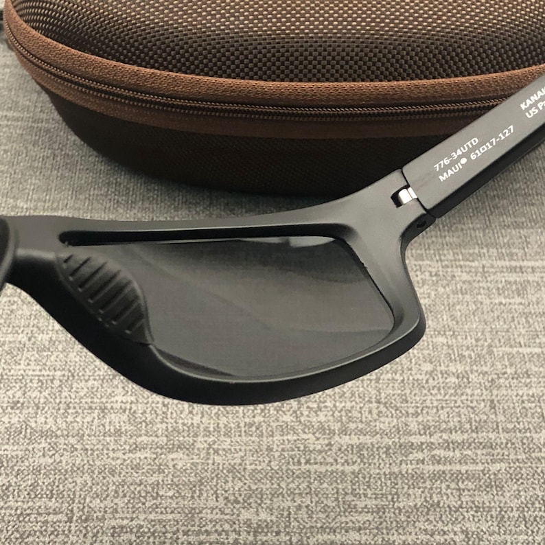 Puede incluir: Gafas de sol negras con lentes oscuras y acabado mate. Las gafas tienen forma rectangular y un agarre texturizado en el lateral. El estuche es marr&oacute;n con cremallera.
