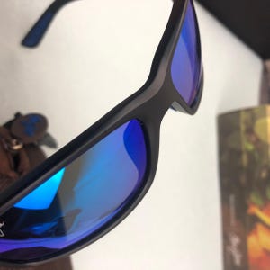May include: Black sunglasses with blue mirrored lenses. The sunglasses have a sleek, modern design with a matte black frame. The lenses reflect a vibrant blue hue, creating a striking contrast.
