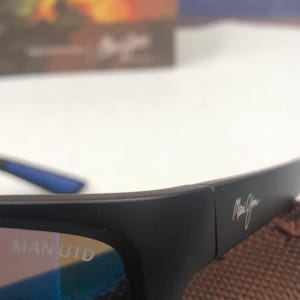 May include: Black sunglasses with the text "MAN UTD" on the frame. The sunglasses have a sleek, modern design with a signature logo on the temple. The lenses appear to have a blue tint.