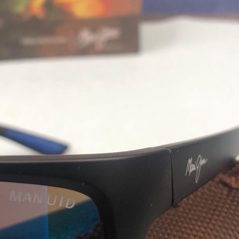 May include: Black sunglasses with the text "MAN UTD" on the frame. The sunglasses have a sleek, modern design with a signature logo on the temple. The lenses appear to have a blue tint.