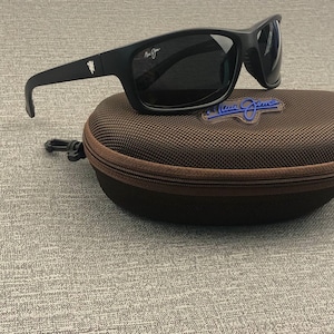 Puede incluir: Gafas de sol negras con lentes oscuras sobre un estuche marr&oacute;n con cremallera. Las gafas tienen una montura rectangular y patillas curvas. El estuche tiene un logotipo azul. Un accesorio elegante.