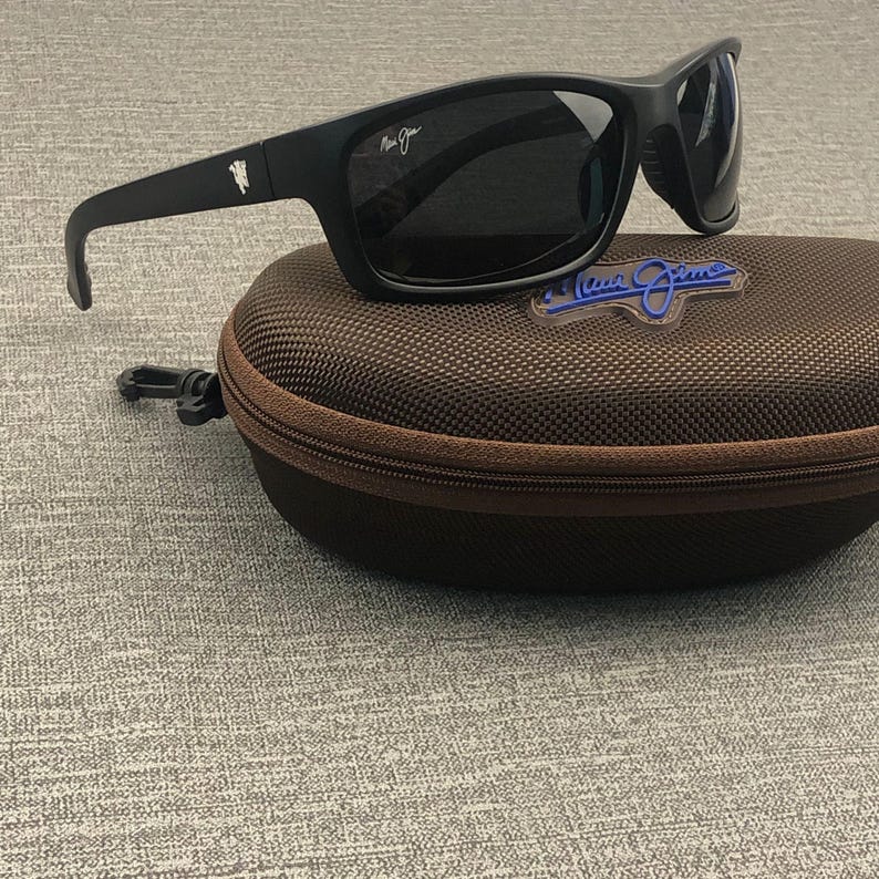 Puede incluir: Gafas de sol negras con lentes oscuras sobre un estuche marr&oacute;n con cremallera. Las gafas tienen una montura rectangular y patillas curvas. El estuche tiene un logotipo azul. Un accesorio elegante.