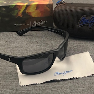 Puede incluir: Gafas de sol negras con lentes oscuras, un pa&ntilde;o de limpieza y un estuche. Las gafas tienen una montura rectangular y la marca "Maui Jim" es visible en las gafas, el pa&ntilde;o y el estuche. La caja en el fondo dice "Welcome to color."
