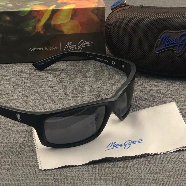Puede incluir: Gafas de sol negras con lentes oscuras, un pa&ntilde;o de limpieza y un estuche. Las gafas tienen una montura rectangular y la marca "Maui Jim" es visible en las gafas, el pa&ntilde;o y el estuche. La caja en el fondo dice "Welcome to color."
