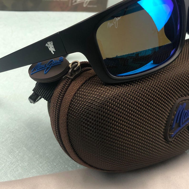 May include: Black sunglasses with blue mirrored lenses rest on a brown zippered case. The sunglasses have a black frame and the brand name is visible on the arm. The case has a logo with the brand name.
