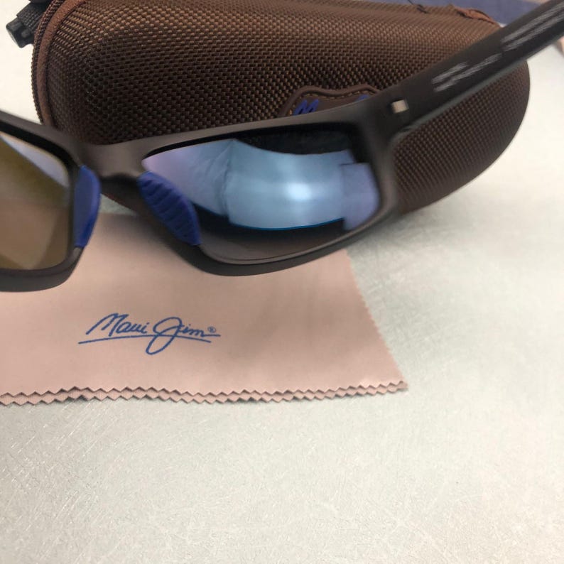 May include: Dark brown sunglasses with blue lenses and a matching case. The sunglasses rest on a pink cleaning cloth with the "Maui Jim" logo in blue script. The lenses reflect a blue hue.