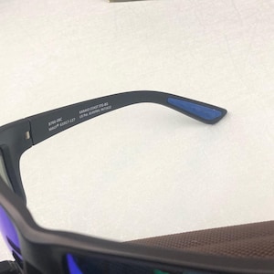 May include: Close-up of black sunglasses with blue lenses and a brown interior. The arm of the glasses has the text "8766-08C MAUI 63017-127" and "KANAIO COAST STG-BG US Pat. 6345986, 5671622".