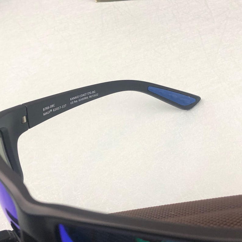 May include: Close-up of black sunglasses with blue lenses and a brown interior. The arm of the glasses has the text "8766-08C MAUI 63017-127" and "KANAIO COAST STG-BG US Pat. 6345986, 5671622".