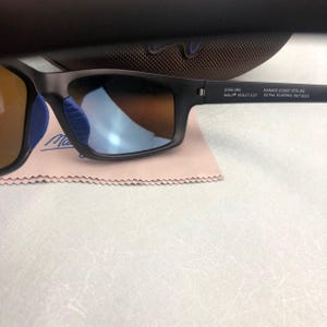 May include: Black rectangular sunglasses with blue nose pads and mirrored lenses. The sunglasses are resting on a pink cleaning cloth. The text on the arm reads "8766-08C MAUI 61017-127 KANAIO COAST STG-BG US Pat. 6145964, 9671622".