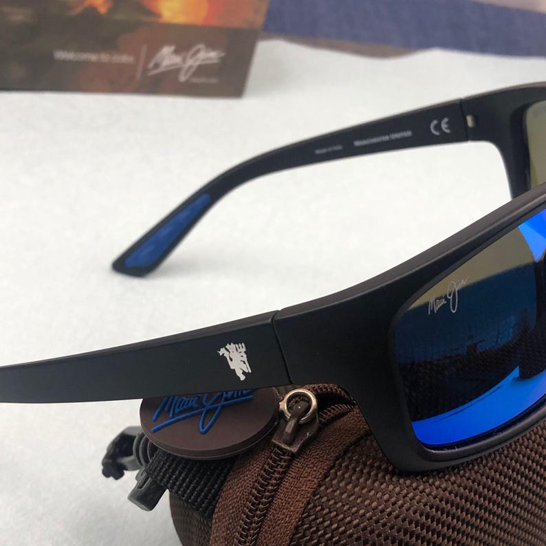 May include: Black sunglasses with blue reflective lenses. The sunglasses have a rectangular frame and a logo on the temple. The sunglasses are resting on a brown case with a zipper.