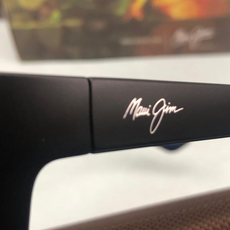 May include: Close-up of black sunglasses with the silver text "Maui Jim" on the temple. The sunglasses are set against a blurred background.