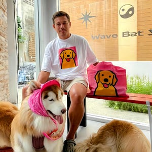 Könnte beinhalten: Weißes T-Shirt und rosa Tragetasche mit einer Cartoon-Hunde-Illustration. Der Hund hat einen gelben Körper, einen schwarzen Umriss und einen lächelnden Ausdruck. Die Tragetasche ist rosa und das T-Shirt ist weiß.