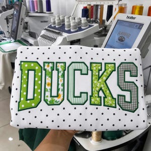 Könnte beinhalten: Ein weißes Sweatshirt mit schwarzem Punktemuster und dem Wort DUCKS als große Applikation in Blockbuchstaben. Jeder Buchstabe zeigt ein anderes Stoffmuster, darunter grüne Punkte, Blumenmuster, grüne Streifen und ein graues Vichy-Karo.