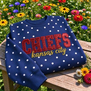 May include: A navy blue crewneck sweatshirt featuring a white polka dot pattern throughout. The front displays the word CHIEFS in red glittery block lettering with the words kansas city embroidered in yellow cursive script below it.