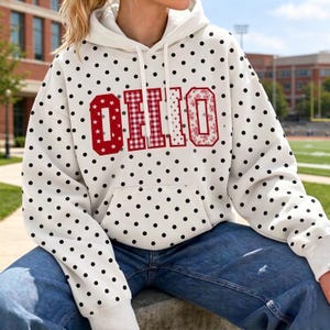 May include: A white hooded sweatshirt featuring a black polka dot pattern throughout. The front displays the word OHIO in large, red-outlined block letters, with each letter filled with a different pattern including stars, gingham, and floral motifs.