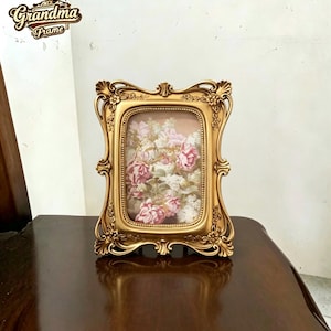 Pode incluir: Uma moldura retangular de cor dourada com detalhes ornamentais em relevo de flores e volutas nas bordas. A moldura contém uma imagem de rosas cor-de-rosa e flores brancas. O texto "Grandma Frame" é visível no canto superior esquerdo.