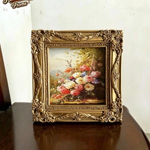 Può includere: Una cornice quadrata con una finitura dorata ornata e in rilievo caratterizzata da dettagli floreali e volute. La cornice contiene un dipinto colorato di un bouquet di rose nelle tonalità del rosso, rosa, giallo e bianco su sfondo verde.
