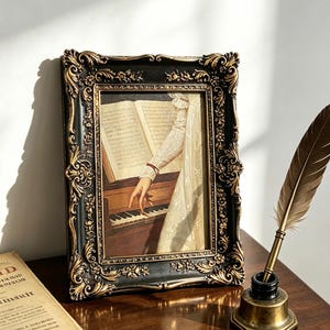 May include: A rectangular picture frame with a black and gold ornate floral border containing a print of a person playing a piano with sheet music. Beside the frame sits a brass inkwell with a brown feather quill and a vintage paper document.