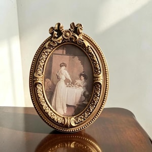 Può includere: Una cornice portafoto ovale color oro con volute in rilievo e un fiocco decorativo sulla parte superiore. La cornice contiene una stampa di due figure in abiti d'epoca vicino a un tavolo, inserita all'interno di un bordo dettagliato.