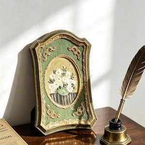 May include: A green and gold ornate picture frame containing a painting of white flowers in a vase, positioned next to a brass inkwell with a brown feather quill pen. The frame features intricate scrollwork details and a central oval display.