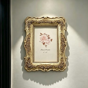 Pode incluir: Uma moldura retangular dourada para fotos com um padrão floral ornamentado em relevo ao longo da borda. A moldura contém uma inserção branca com uma ilustração de rosa rosa e o texto impresso "Photo Frame 4" x 6"". A moldura está montada.