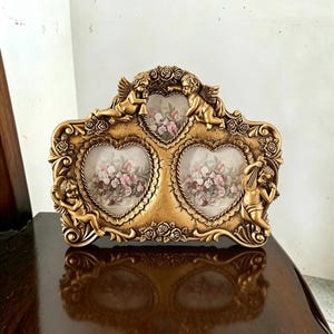 May include: A gold-colored triple photo frame featuring three heart-shaped openings. The frame is decorated with sculpted cherubs, floral rose motifs, and intricate scrollwork. Each opening contains a print of pink and white flowers.