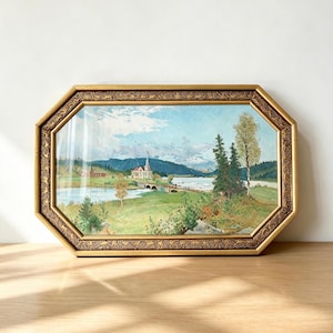 May include: An octagonal framed painting depicting a landscape with a river, a bridge, and a building with a steeple. The frame is gold-colored with a dark, intricate, embossed pattern along the border. The scene features green fields, trees, and mountains under a blue sky with white clouds.