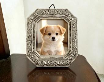 May include: A small, light tan and white puppy with floppy ears and dark eyes is centered within an ornate, silver-toned octagonal picture frame. The frame features intricate embossed floral and scrollwork detailing along its wide border.