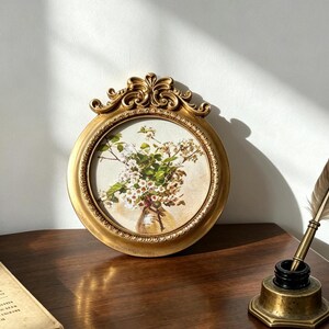 May include: A circular gold-toned frame with ornate scrollwork at the top, containing a print of white and green flowers in a glass vase. The frame features a beaded inner border and a smooth outer edge, showcasing a detailed floral illustration.