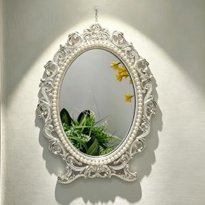 May include: An oval wall mirror featuring a silver-toned frame with intricate scrollwork and a border of small white pearls. The mirror reflects green foliage and yellow flowers, and it is suspended by a small metal chain from a wall hook.