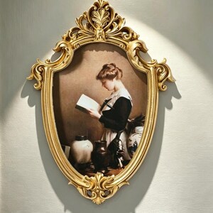 May include: An oval wall mirror featuring an ornate gold frame with intricate scrollwork and floral carvings. The mirror displays a print of a person in a black and white outfit reading a book, with several ceramic vases placed on a table nearby.