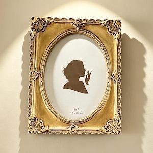 May include: A rectangular gold picture frame with ornate, scalloped edges and decorative corner detailing. The frame features an oval inner border and contains a white insert with a brown silhouette of a person and the text 5x7 13x18cm.