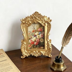 May include: An ornate gold-colored picture frame with intricate scrollwork detailing, containing a print of a colorful floral bouquet. Beside the frame sits a metallic inkwell with a brown feather quill pen and a piece of aged paper with text.