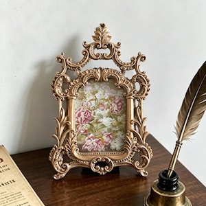 Peut inclure: Un cadre photo doré orné de volutes et de motifs de feuilles complexes, contenant une impression florale de roses roses et de fleurs blanches. À côté se trouve un encrier en laiton avec une plume brune et un morceau de papier vieilli.