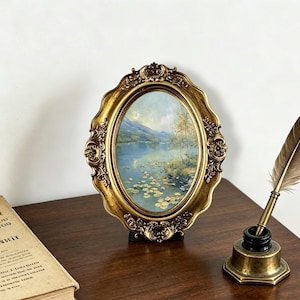 Puede incluir: Un marco de fotos ovalado con un acabado dorado ornamentado que contiene una pintura de paisaje de un lago con nenúfares y montañas. Junto al marco hay un tintero de latón con una pluma marrón y un libro antiguo con texto visible.