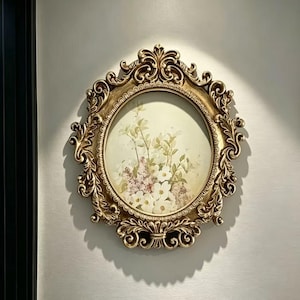 Puede incluir: Un marco de pared ovalado con un borde de resina dorado ornamentado, con volutas intrincadas y tallas en forma de hoja. El centro contiene una impresión de tonos claros que representa un ramo de flores blancas y moradas pálidas.