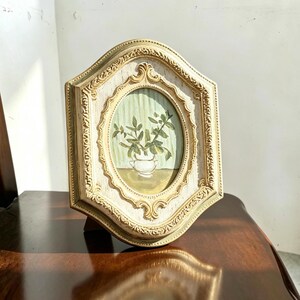 May include: An ornate, hexagonal-shaped picture frame with a distressed cream and gold finish. The frame holds an oval painting depicting a small white vase containing green leafy stems against a pale green, vertically striped background.