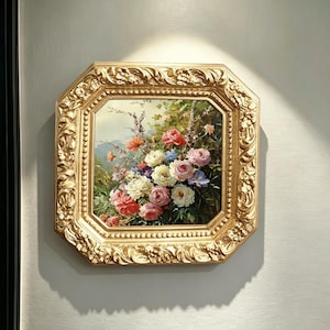May include: A square painting of a floral bouquet featuring pink, white, and blue roses with green foliage, set within an ornate, thick gold-colored frame with intricate carved detailing and a beaded inner border, mounted on a plain wall.