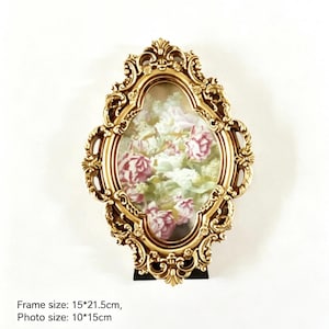 Pode incluir: Uma moldura oval de cor dourada com detalhes ornamentados em relevo. A moldura contém uma estampa floral com rosas cor-de-rosa e flores brancas. A moldura mede 15 por 21,5 cm, com uma área de exibição de foto de 10 por 15 cm.