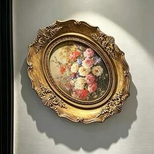 Peut inclure: Un cadre mural ovale avec une finition dorée métallique et des détails en relief ornés. Le cadre entoure une impression florale colorée représentant un bouquet de roses et d'autres fleurs dans des tons de rose, blanc, rouge et bleu sur un fond sombre.