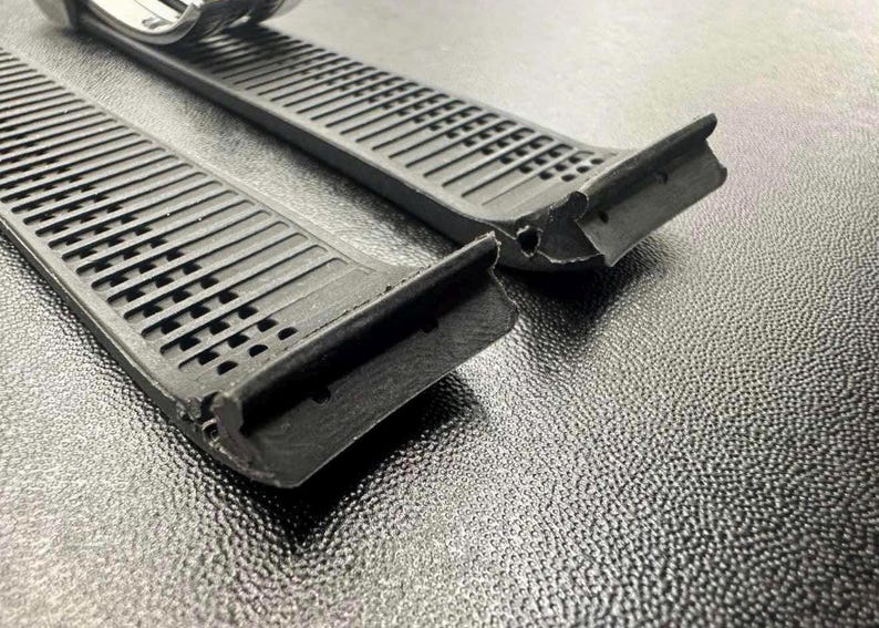 Pu&ograve; includere: Primo piano di due cinturini neri per orologi con un design strutturato e scanalato. I cinturini hanno una serie di ritagli rettangolari e sono realizzati in un materiale flessibile. Il cinturino dell'orologio &egrave; parzialmente visibile sullo sfondo.