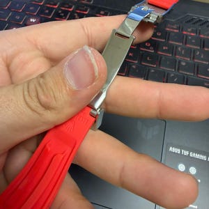 Peut inclure: Un bracelet de montre rouge avec un fermoir argent&eacute; est tenu contre un clavier d'ordinateur portable. Le bracelet est fait d'un mat&eacute;riau souple et pr&eacute;sente un motif textur&eacute;. Le fermoir est brillant et m&eacute;tallique.