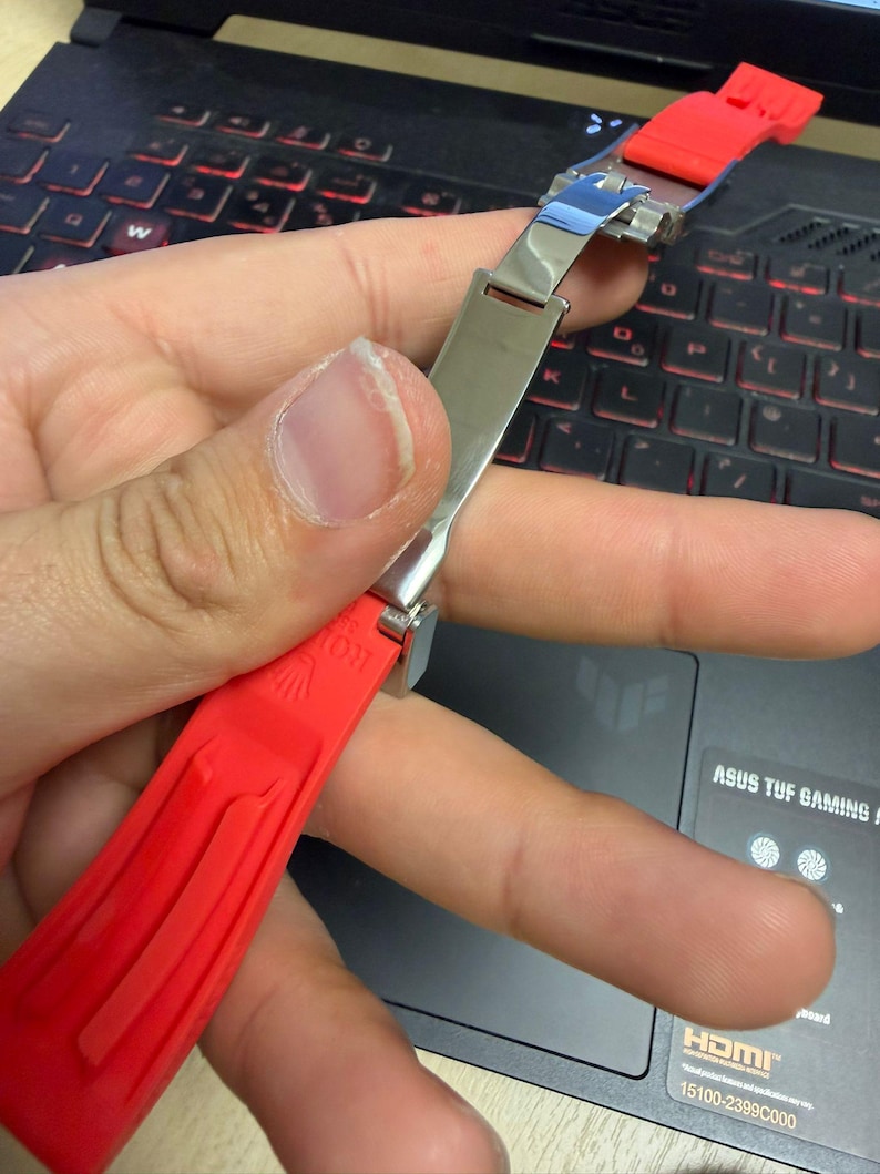 Peut inclure: Un bracelet de montre rouge avec un fermoir argent&eacute; est tenu contre un clavier d'ordinateur portable. Le bracelet est fait d'un mat&eacute;riau souple et pr&eacute;sente un motif textur&eacute;. Le fermoir est brillant et m&eacute;tallique.