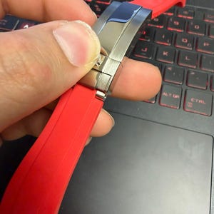 Peut inclure: Un bracelet de montre rouge avec un fermoir en acier inoxydable. Le bracelet est fait d'un mat&eacute;riau souple et est tenu par une main. Le fermoir est argent&eacute; et a une finition polie. L'arri&egrave;re-plan est un ordinateur portable noir.